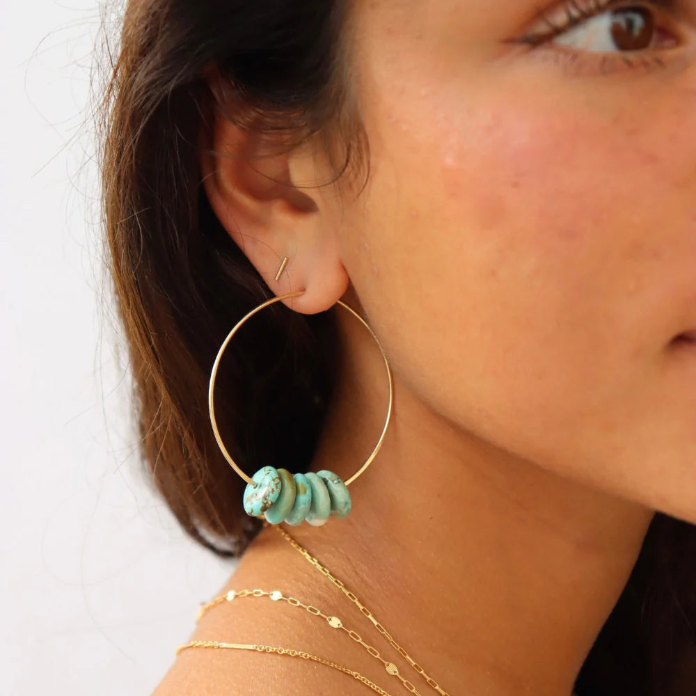 Close-up of a person wearing gold hoop earrings with turquoise beads on a white background